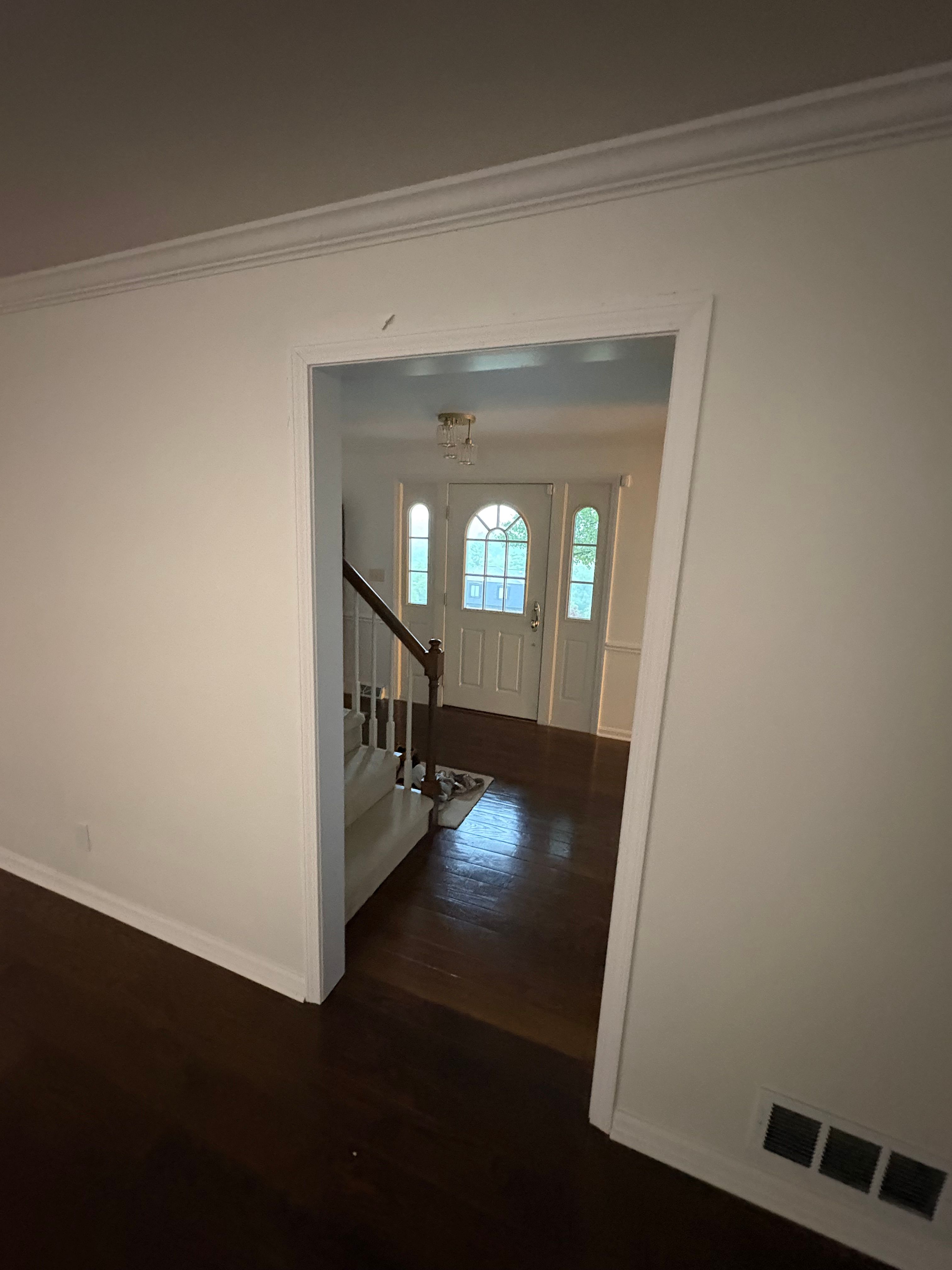 A view through a wide white-cased doorway into an entry hall, where a staircase with white balusters and a dark stained handrail rises on the left and the panelled front door ahead carries an arched window flanked by two tall arched sidelights, all above dark stained hardwood floors.