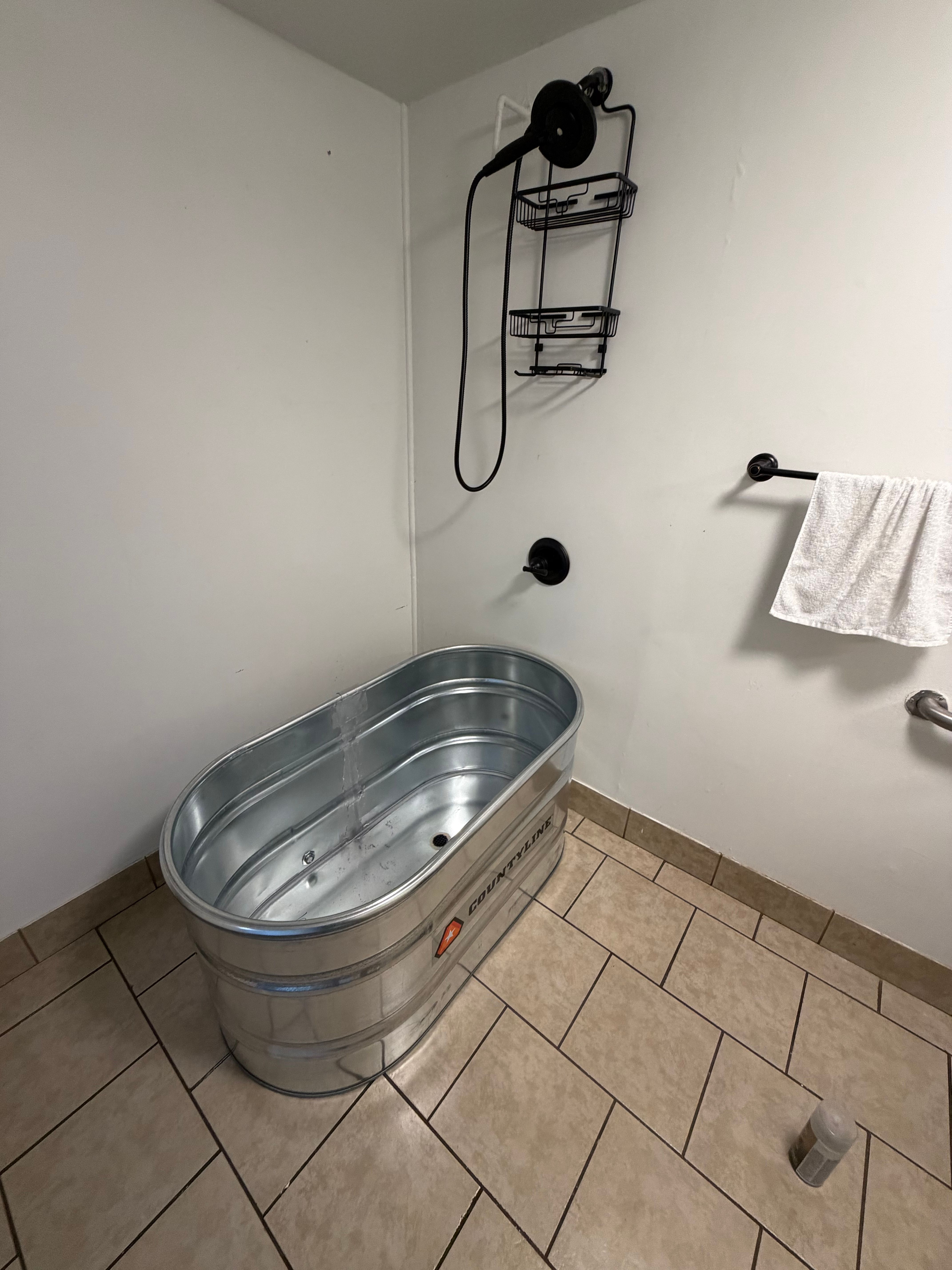 A galvanized stock tank tub on a tile floor in a finished basement bathroom, below a handheld shower on a hose, a shower valve and a towel bar on the wall.