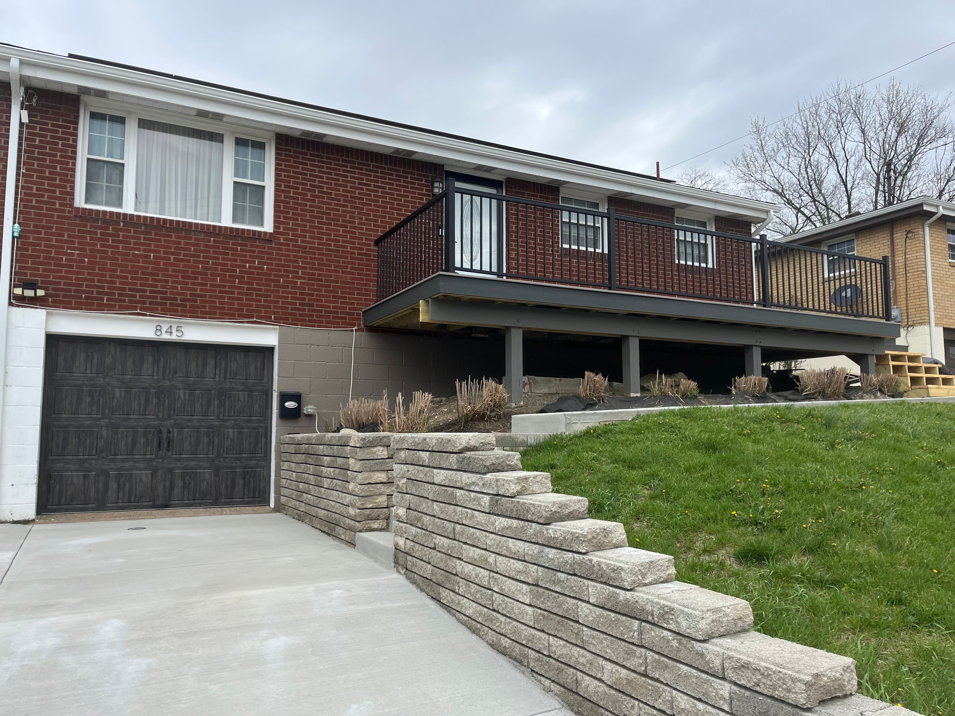 A stacked stone retaining wall and steps running beside a concrete driveway, below a brick house with a raised deck.