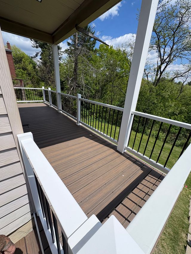 A covered deck under a shingled roof, with tan composite boards framed by a dark brown border, white posts and top rails, and black metal balusters looking out to a lawn and trees.