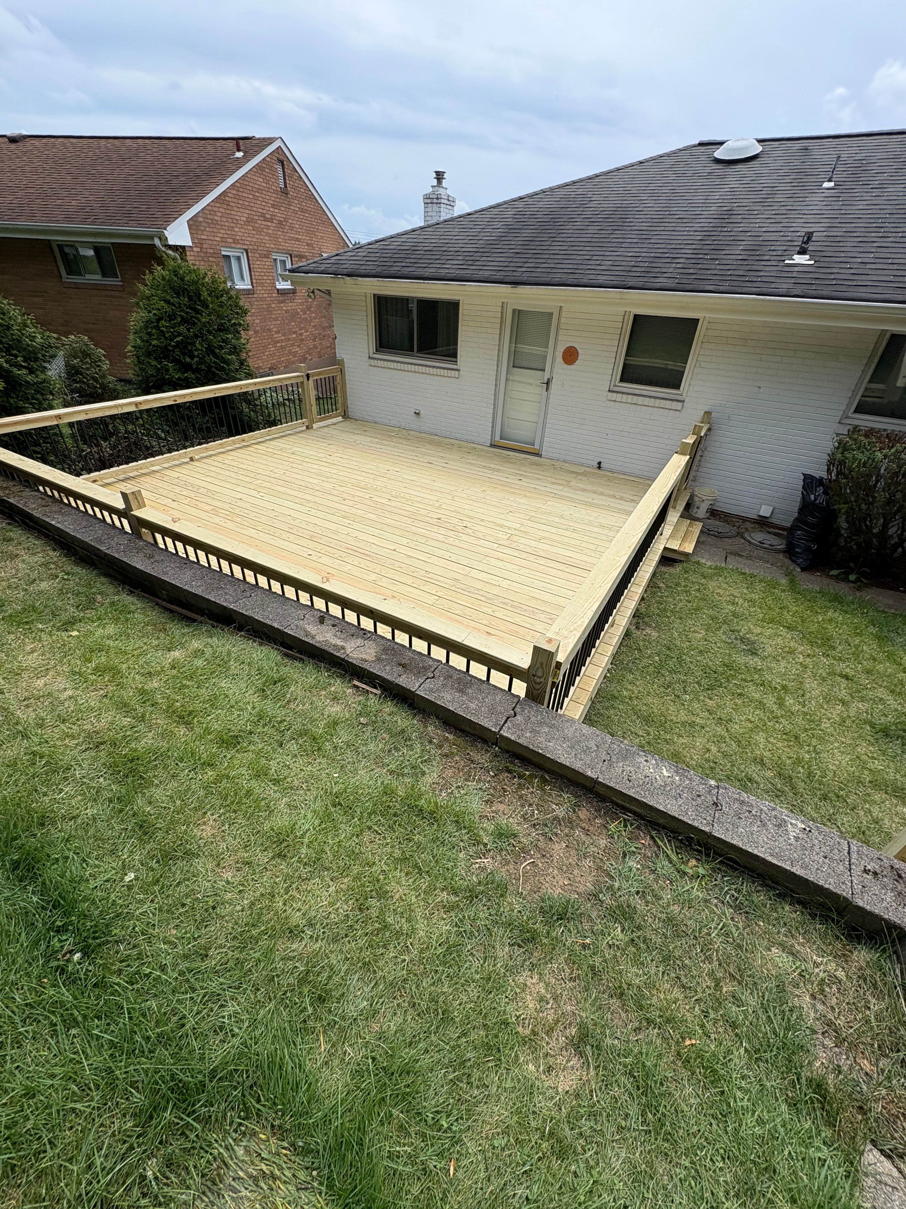 A newly built low pressure-treated pine deck with a matching wood railing and black metal balusters, running along the back of a white brick ranch house above a mown lawn.