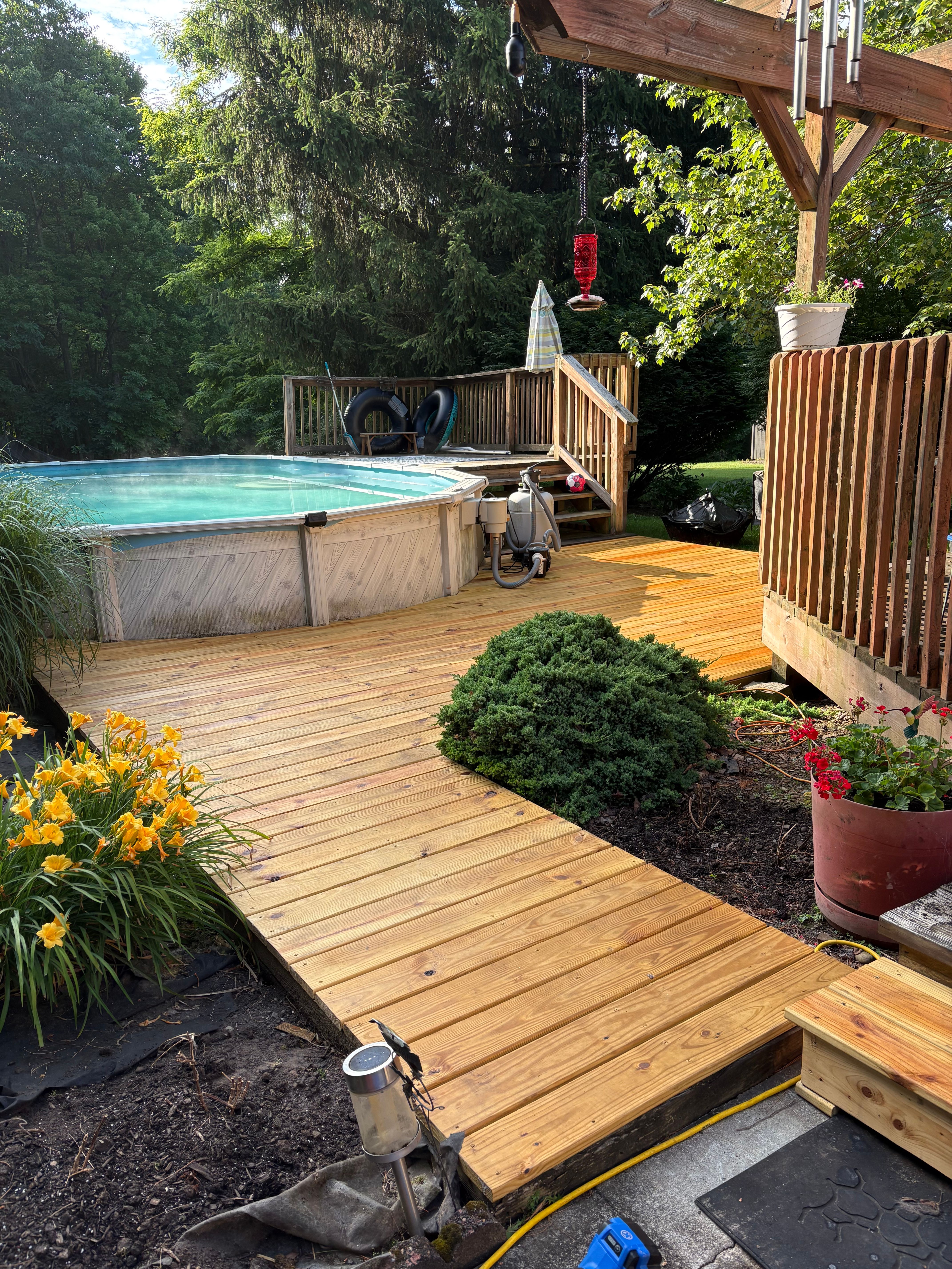 A pressure-treated pine deck wrapping around an above-ground pool, with a railed landing and steps at the pool edge and beds of orange day lilies and low shrubs along the deck's outer edge.
