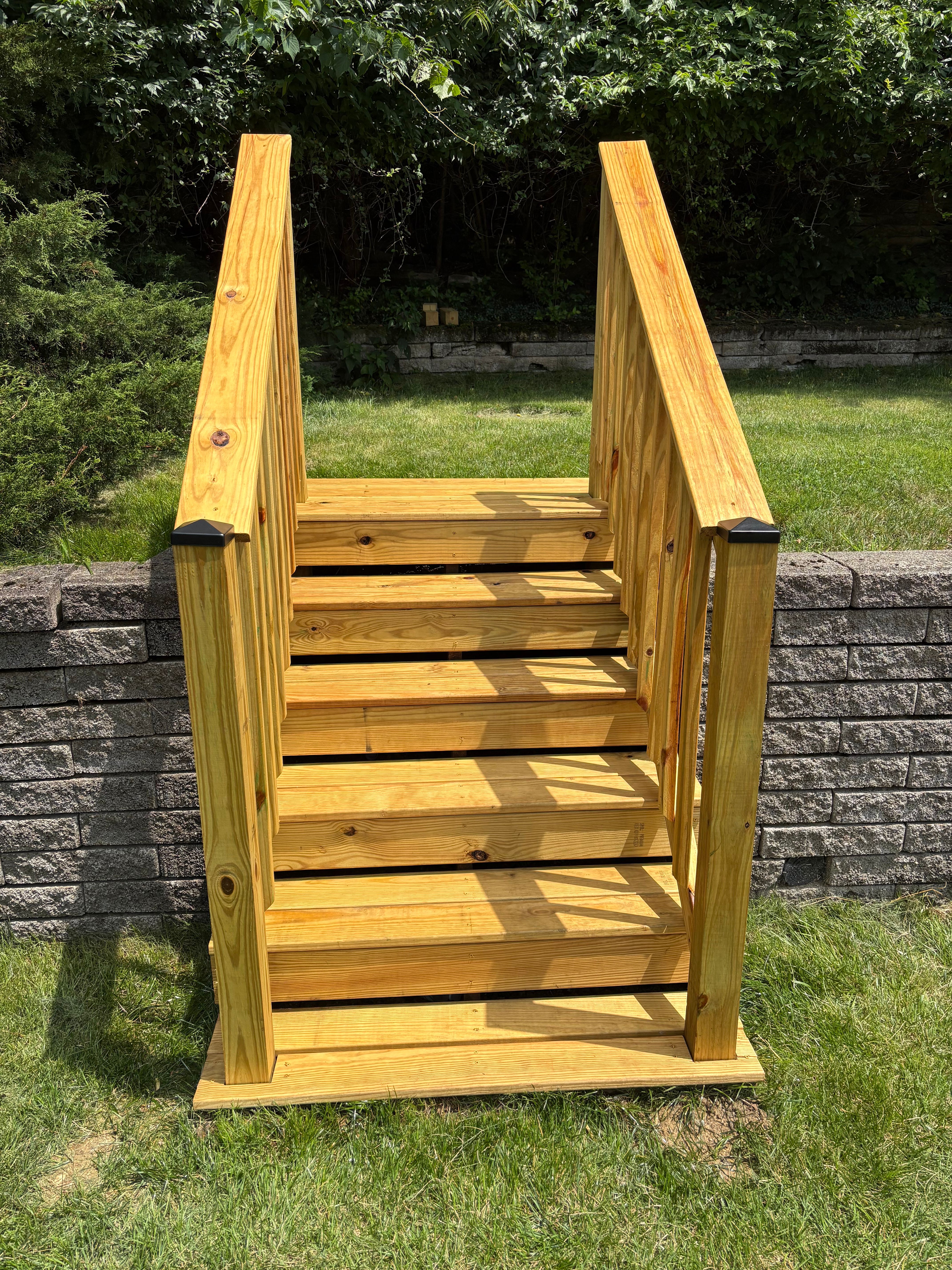 A short flight of newly built pressure-treated pine steps with a railing on both sides and black post caps, set into a stacked grey block retaining wall with a mown lawn behind it.