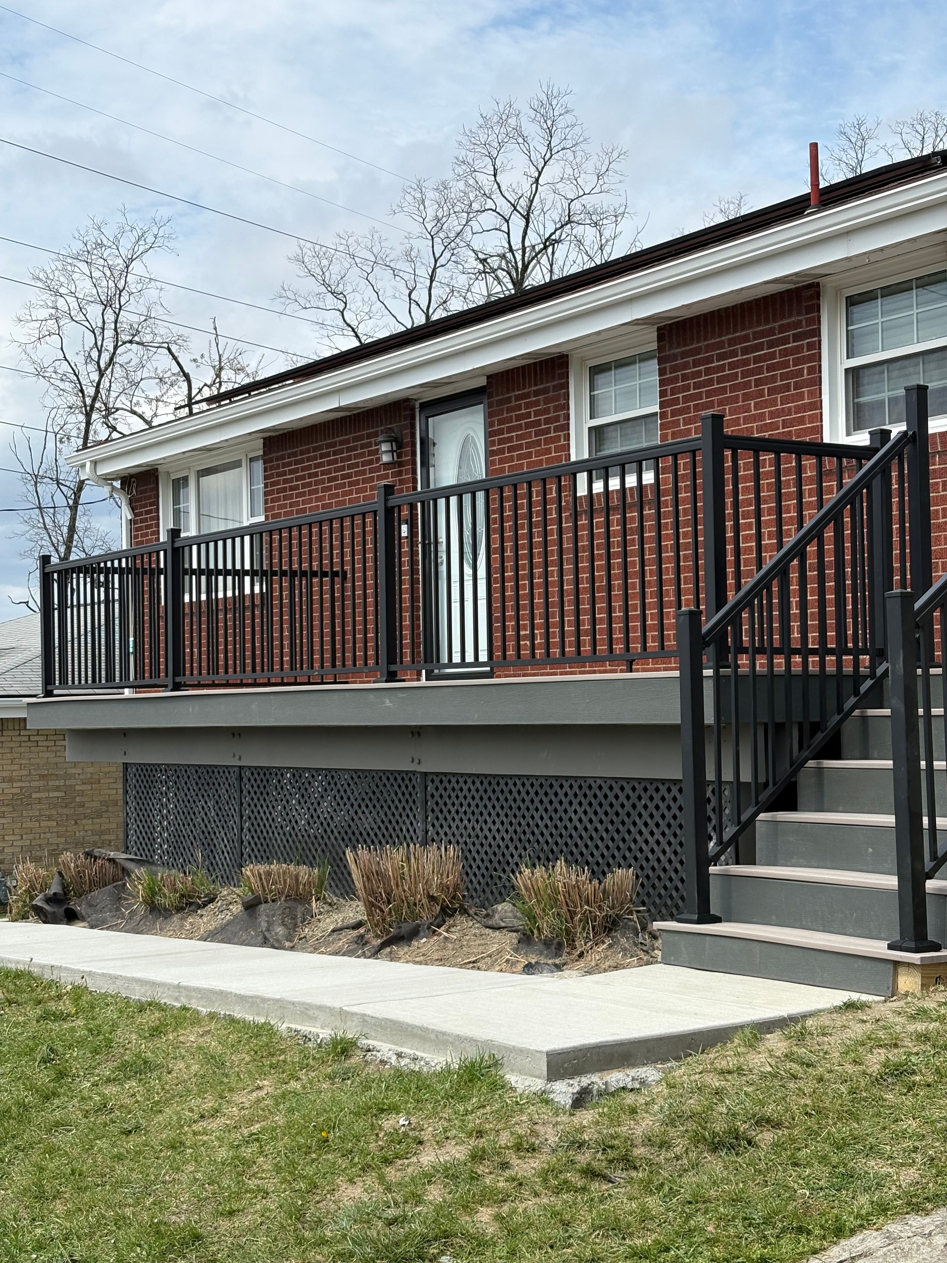 A grey composite deck with a black aluminum railing running across the front of a red brick house, with matching black stair railings to the right and a dark lattice skirt below the deck boards.