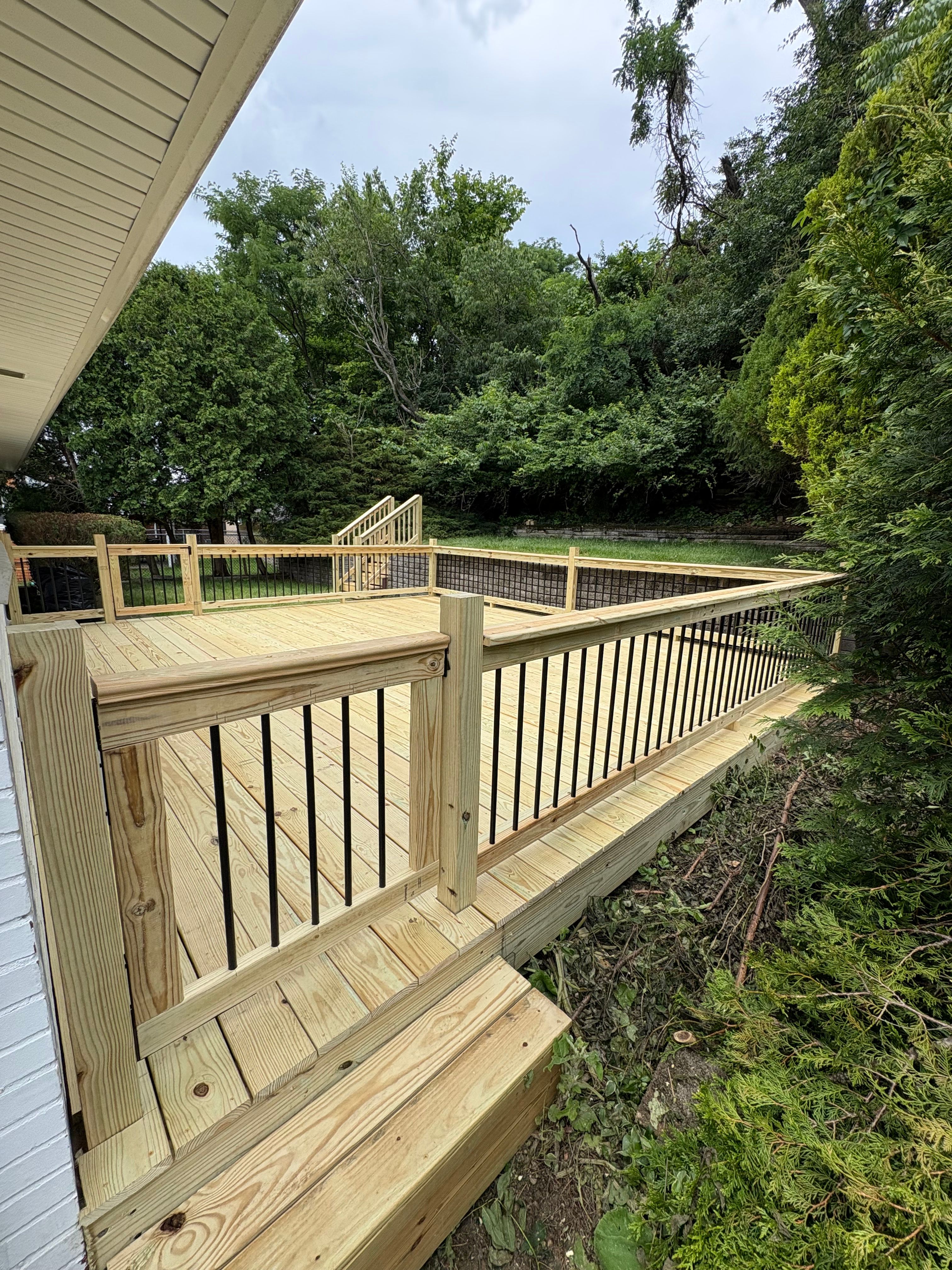 A newly built pressure-treated pine deck with a wood railing and black metal balusters, running alongside a white-sided house with a dense tree line behind it.