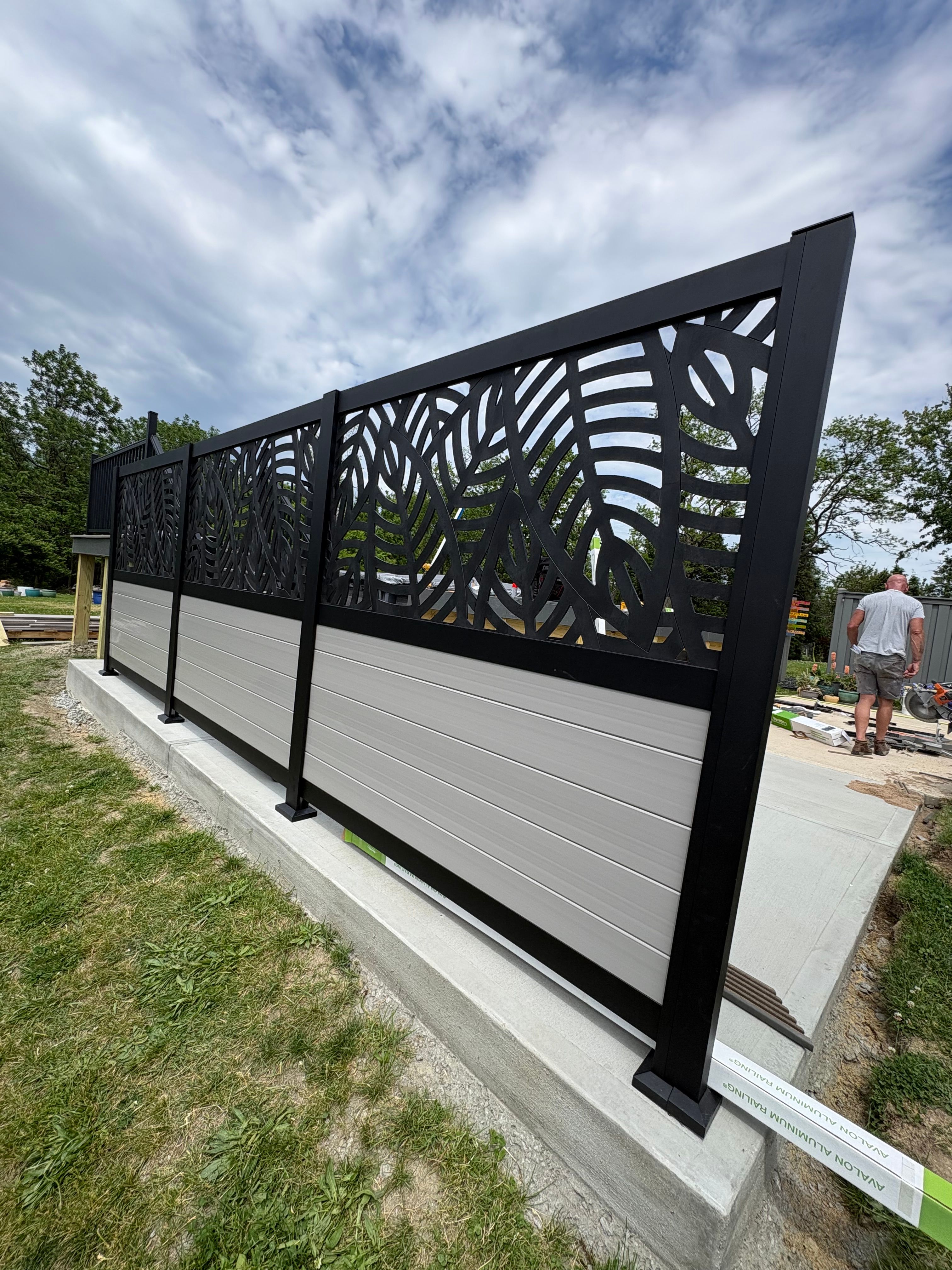 A black aluminum privacy screen with a leaf-pattern cutout above solid panels, set on a new concrete pad beside a lawn.