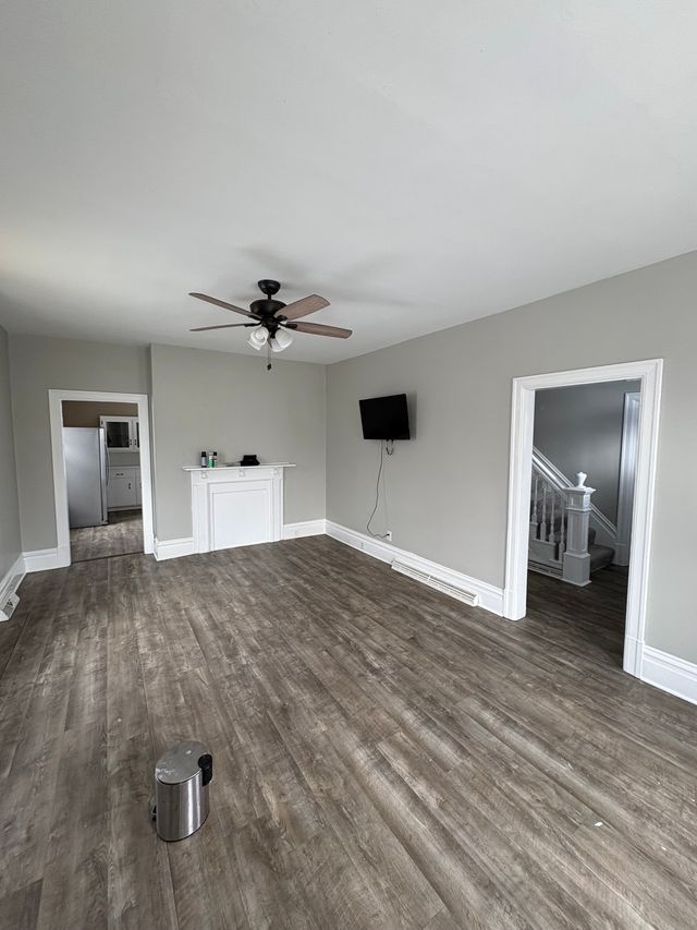 An empty remodeled room with grey walls, white trim and dark wood-look plank flooring, a ceiling fan overhead and a white staircase through the doorway.