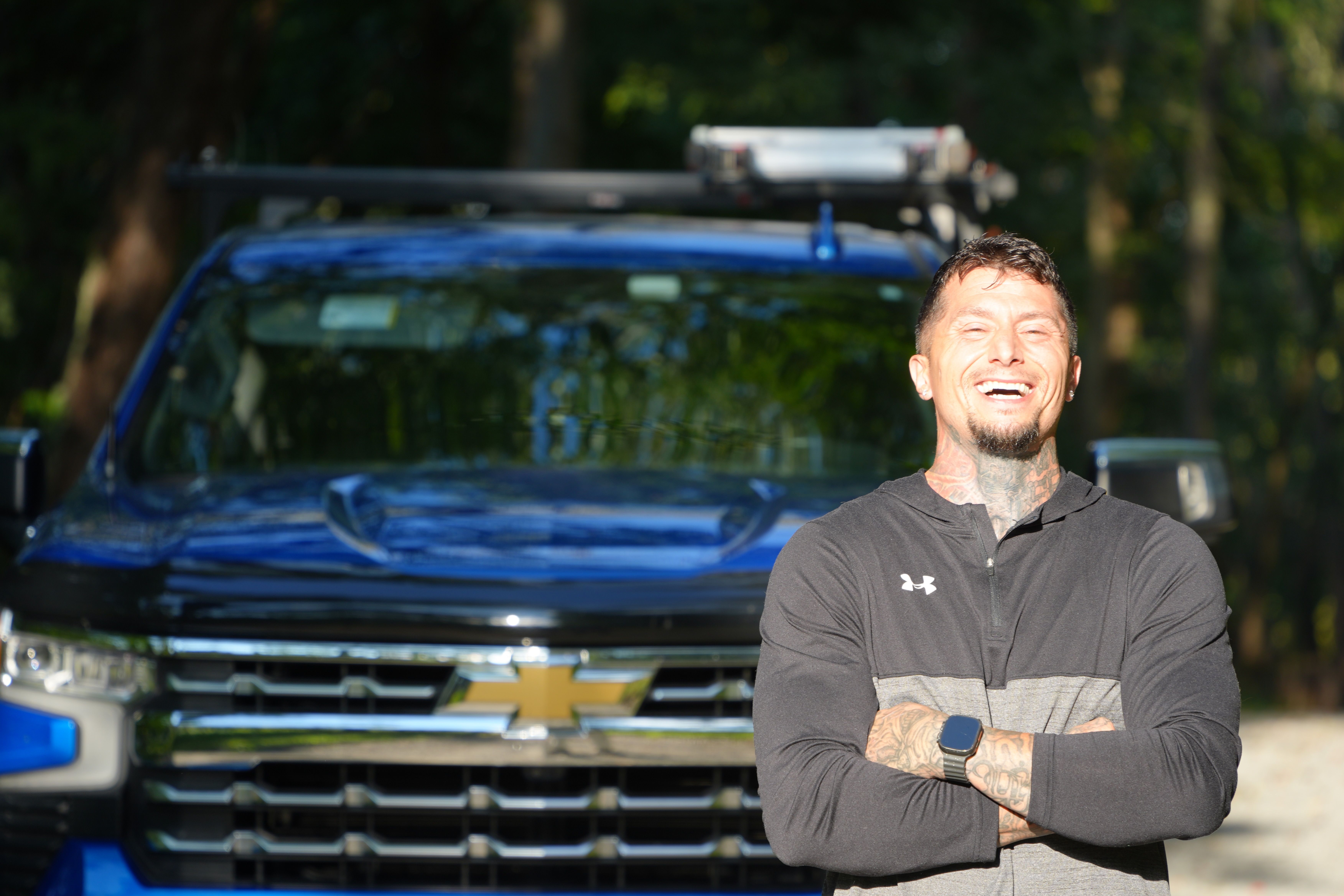 Anthony laughing with his arms crossed in front of a blue Chevrolet pickup