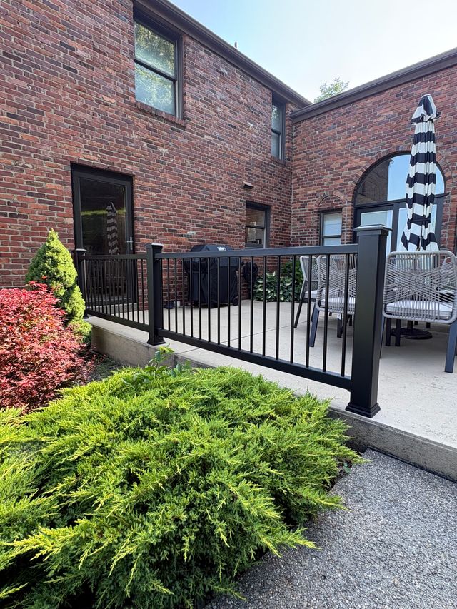 A black metal railing running along the edge of a raised concrete patio beside a red brick house, with cushioned chairs, a folded striped umbrella and low shrubs below.
