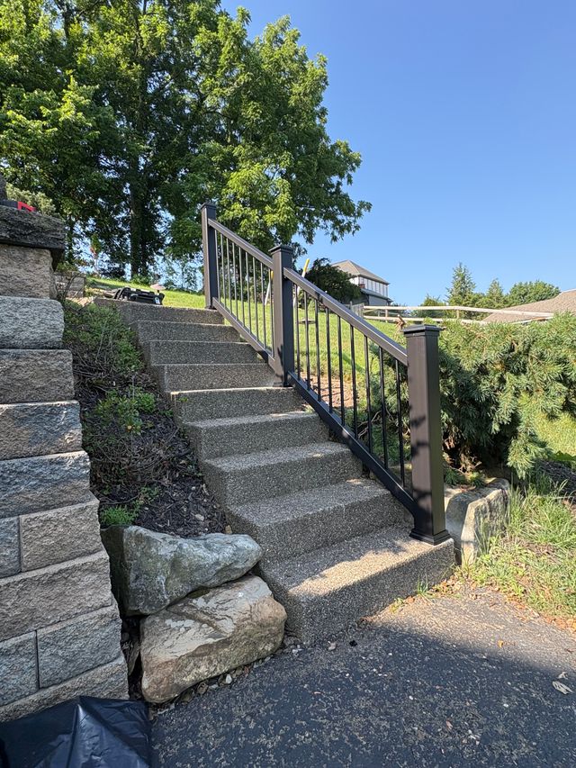 A flight of poured concrete steps climbing a slope from a driveway, with a black metal railing on one side and a grey stacked block wall on the other.