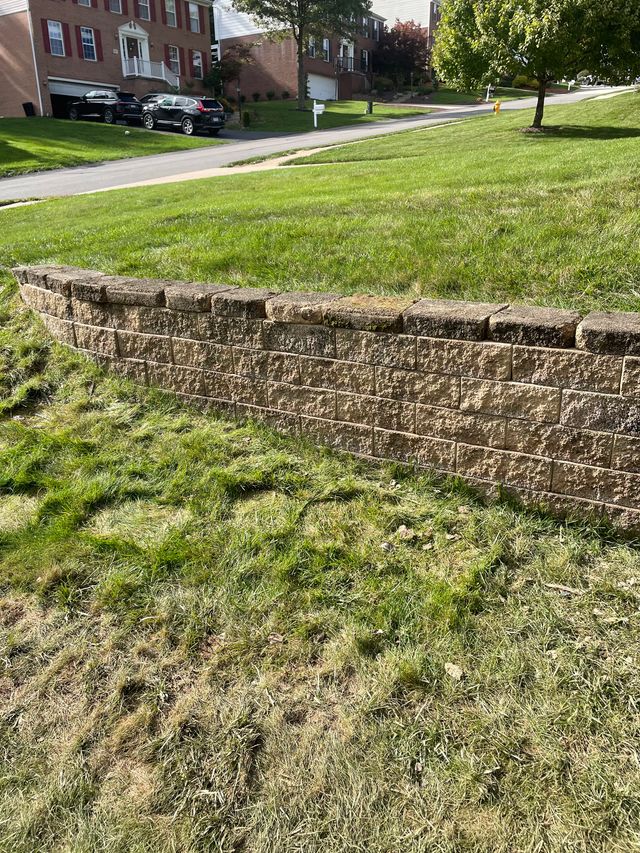 A long tan block retaining wall curving across a mown lawn, capped with a flat top course, with a street and houses beyond.